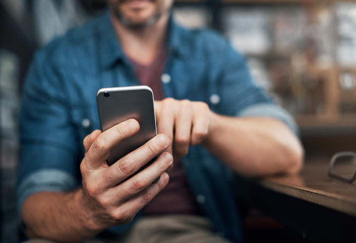 Bearded man in a blue denim shirt sitting at a bar sending a text message.