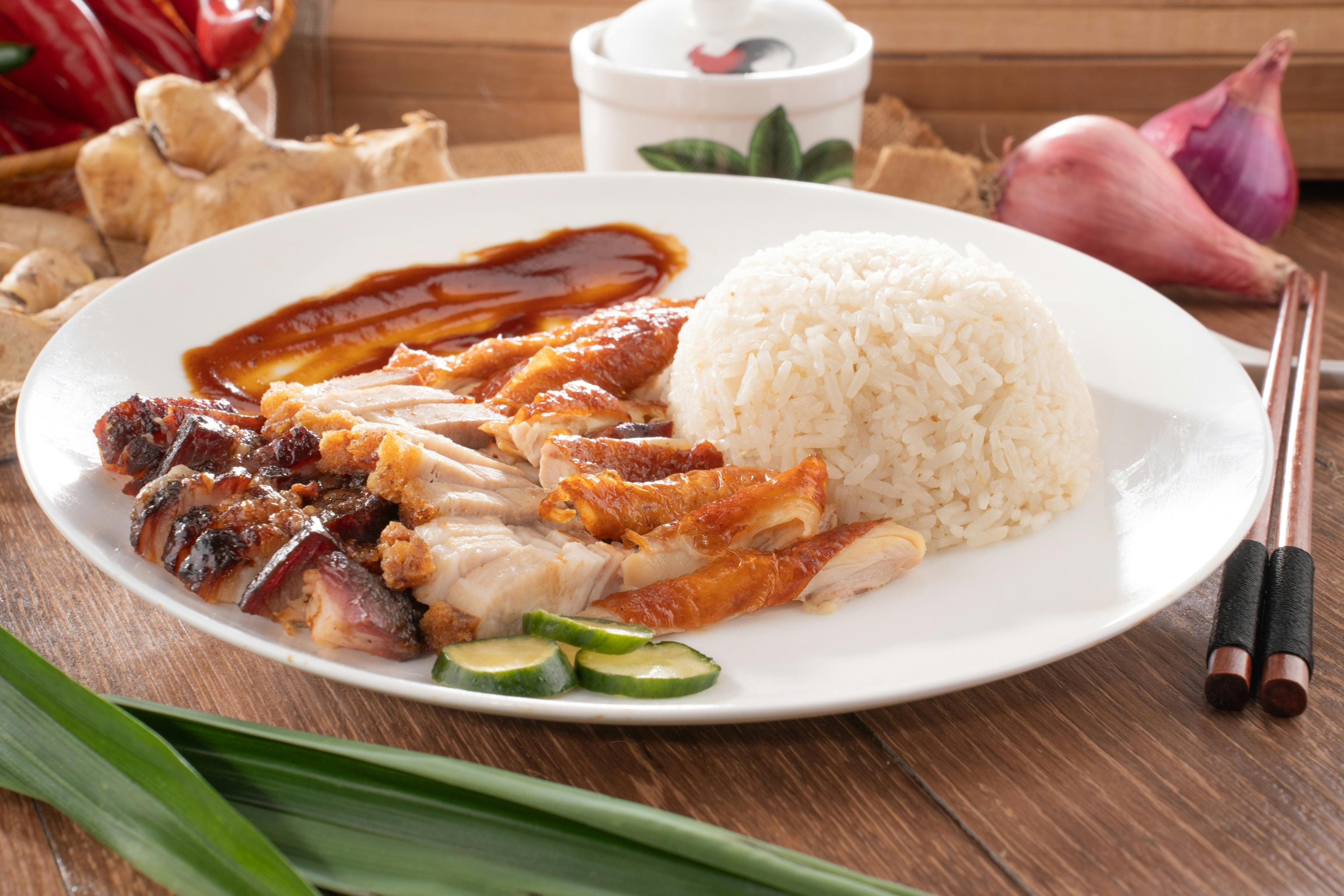 This plate presents a delectable combination of roasted meats, featuring sliced char siu and crispy-skinned roasted chicken served alongside a neat mound of white rice. The dish is finished with a generous smear of savory sauce, fresh cucumber slices, and is set against a rustic background with ginger, red onions, and wooden chopsticks.