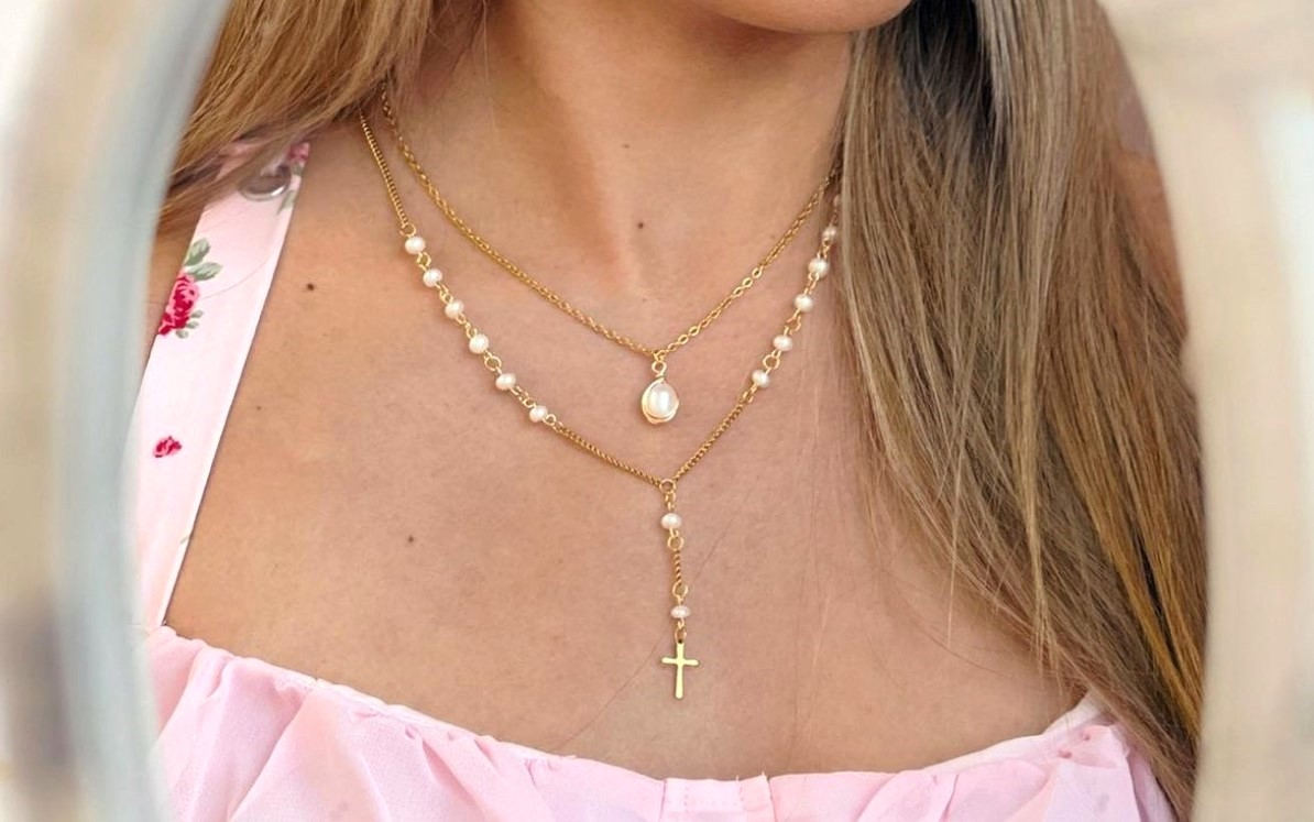 A close-up view of a woman's neck wearing two gold-colored, stainless steel necklaces layered together; one with a single pearl pendant and the other a cross with pearl accents.