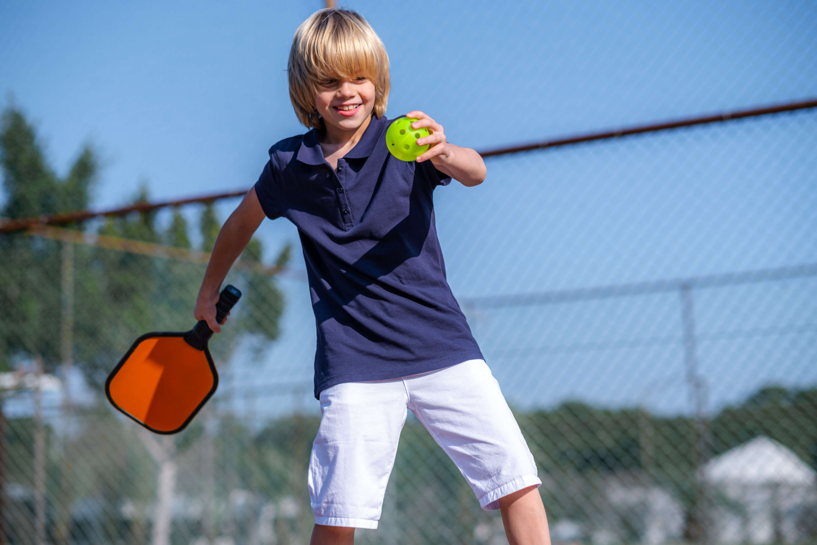 A blonde-haired boy poised to play, holding a pickleball paddle and focusing intently on the game.