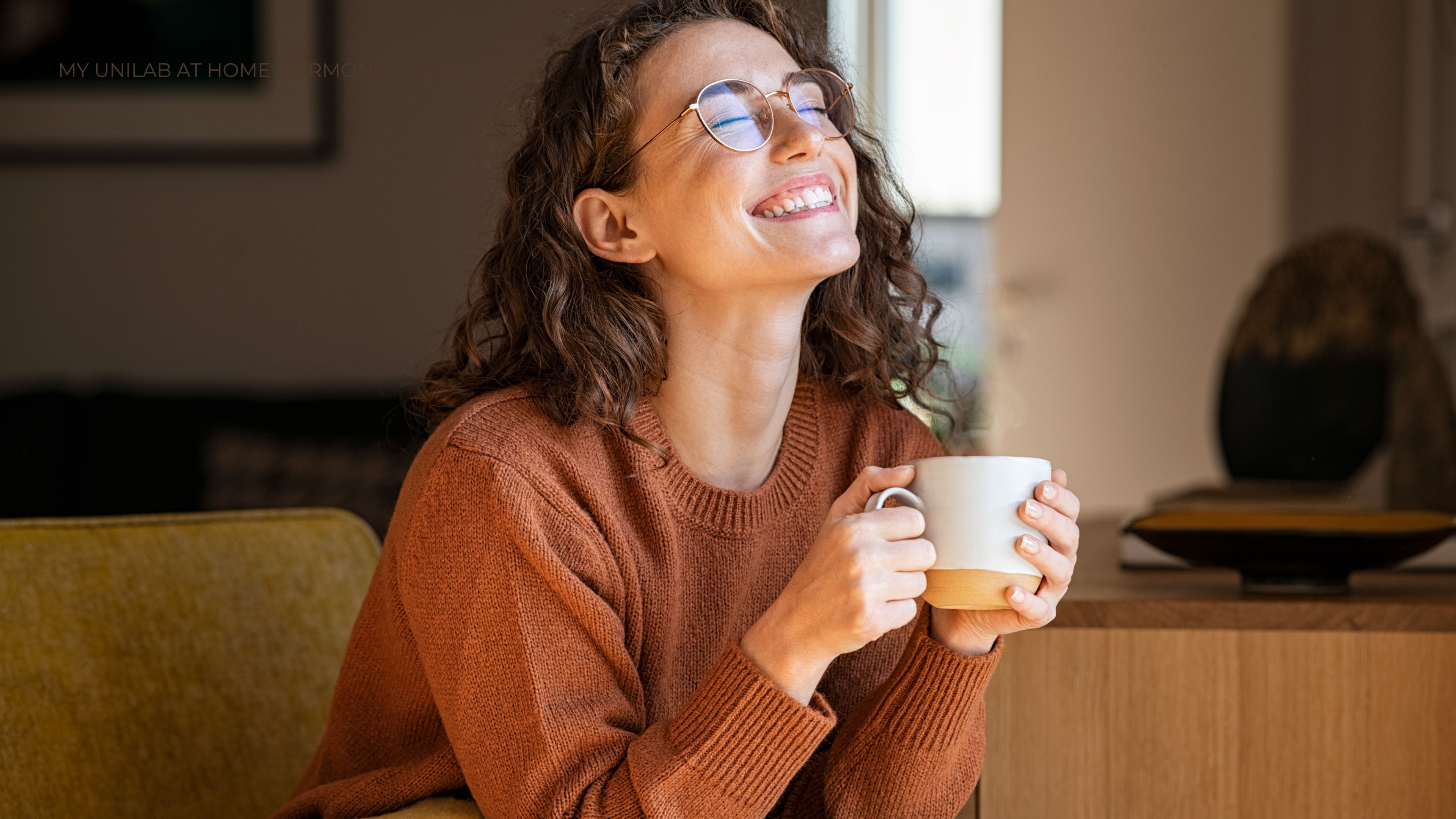 Women looks up towards the ceiling of a room with her eyes closed and a big smile on her face holding coffee.