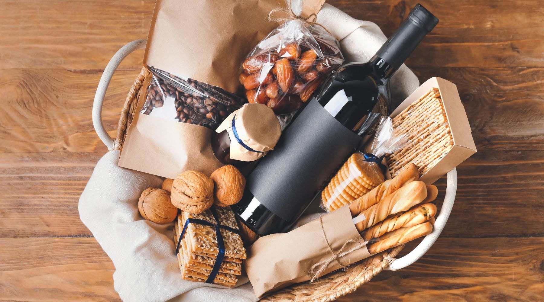 Basket with wine, snacks, and packed goods on wooden surface.