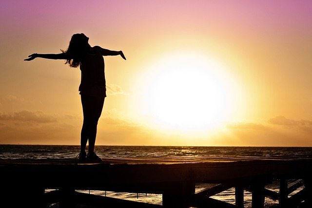 A girl spreading her arms standing on a dock facing the sun next to the ocean.
