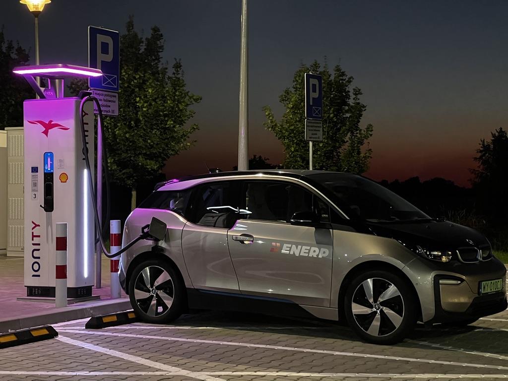 Na zdjęciu widoczny jest nowoczesny dom jednorodzinny, przed którym stoi samochód elektryczny podłączony do wallboxa. Użytkownik pojazdu korzysta z wygodnego ładowania, co wpływa na niskie koszty eksploatacji samochodu elektrycznego.