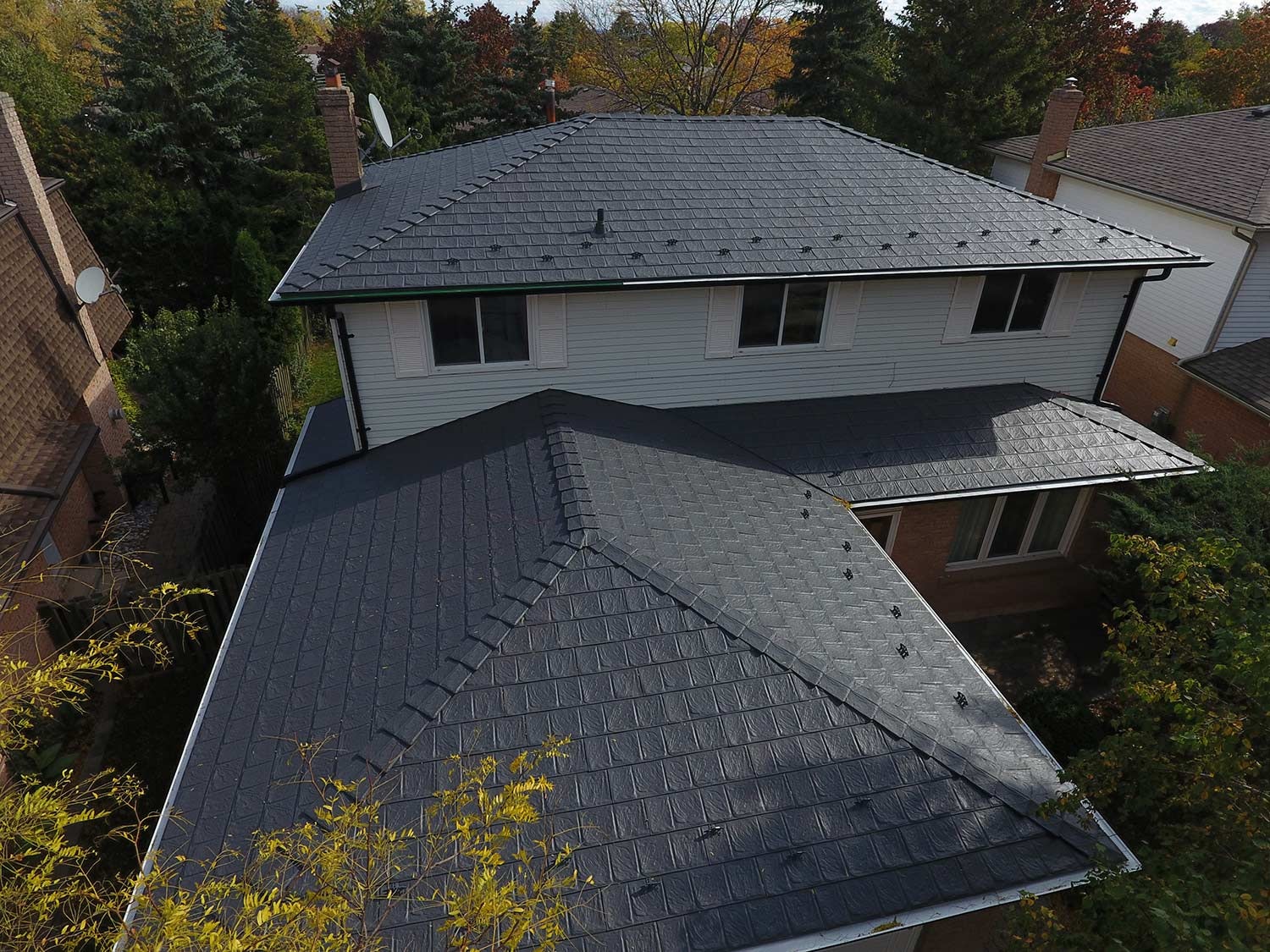 A side view aerial picture of a home with a dark metal roof, surrounded by other homes and trees.