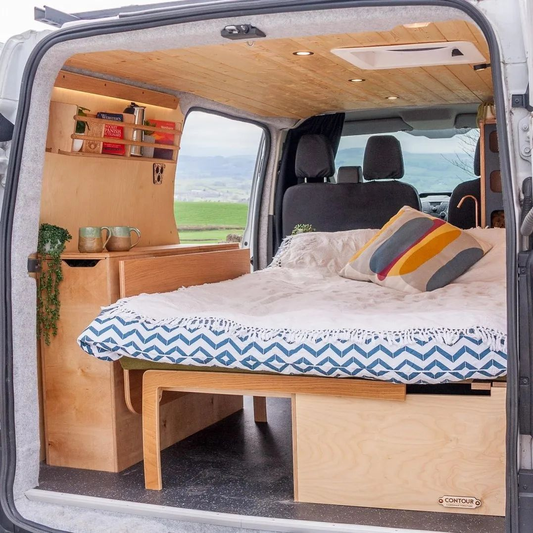 The back of a van conversion with a well made bed next to a table with two drinking cups and a plant. 