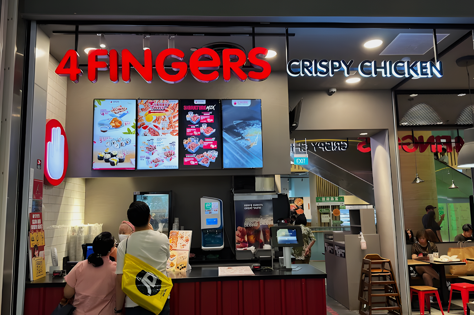 Storefront of 4Fingers Crispy Chicken with a red and white sign. Two customers wait at the counter, illuminated menus above displaying food items.