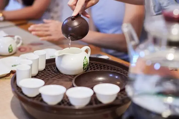 A person pours tea from a small clay pot into a white pitcher on a traditional wooden tea tray. Several small white teacups are arranged in the foreground, creating a warm and social atmosphere.