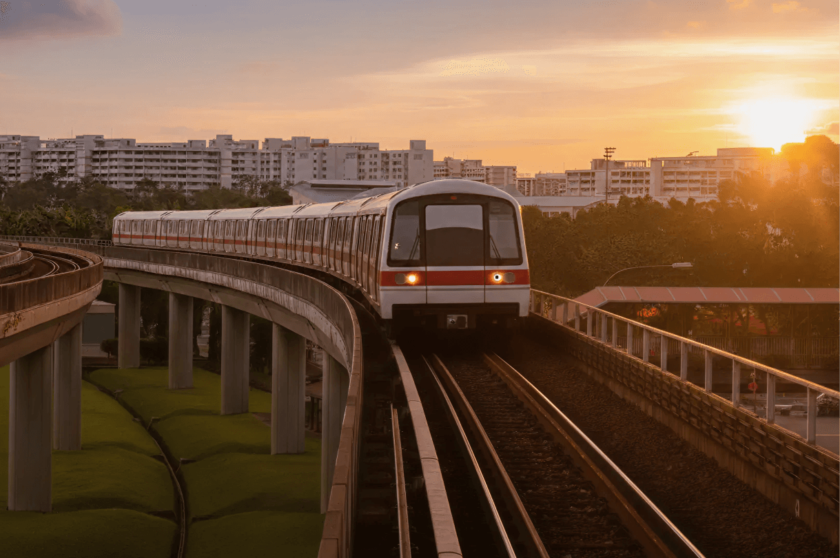 An elevated metro train curves along raised tracks at sunset, with warm golden light illuminating the train, nearby trees, and apartment buildings in the background.
