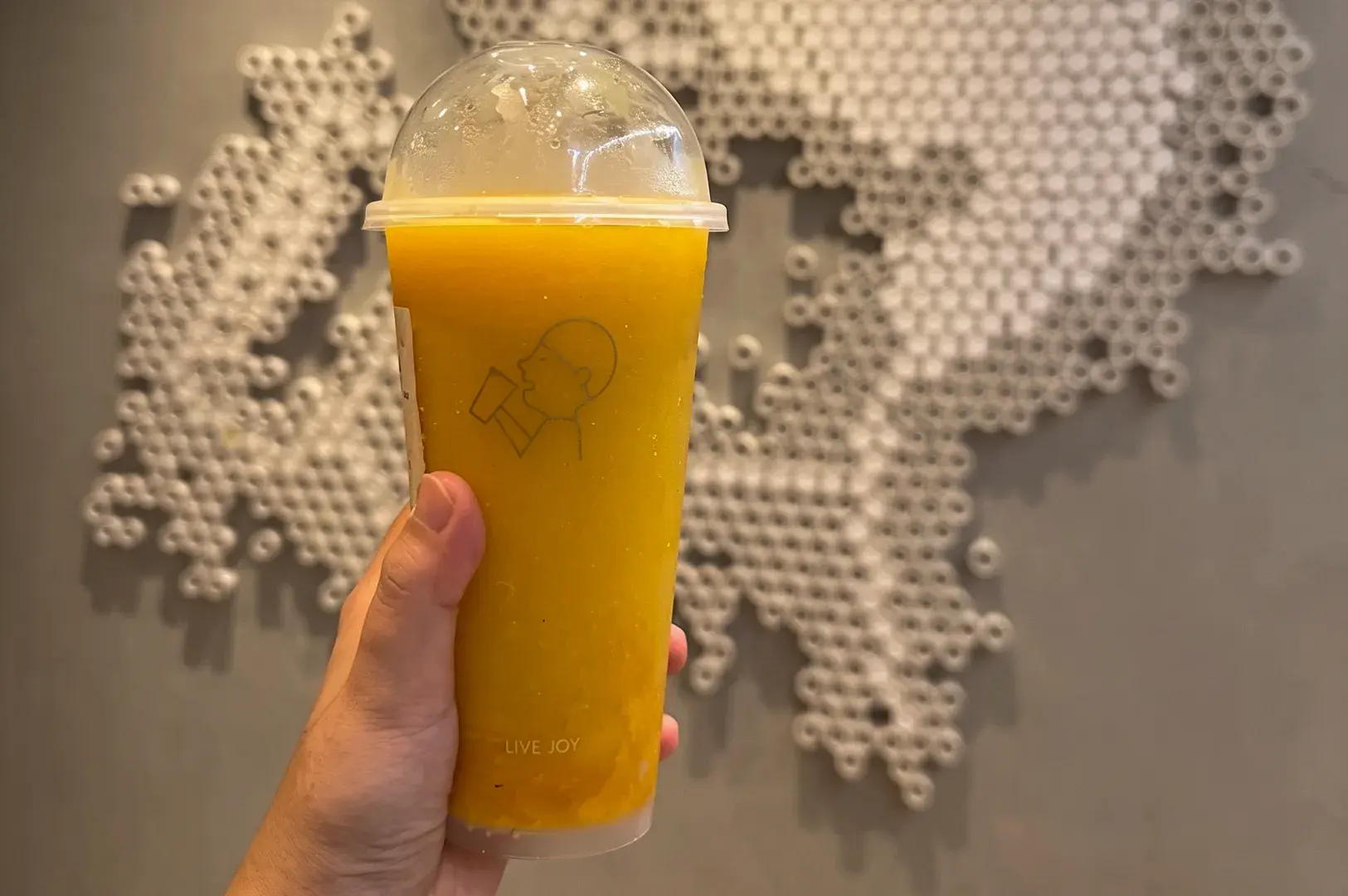Hand holding a branded HEYTEA yellow fruit beverage cup against a textured grey wall with an abstract white circular pattern.