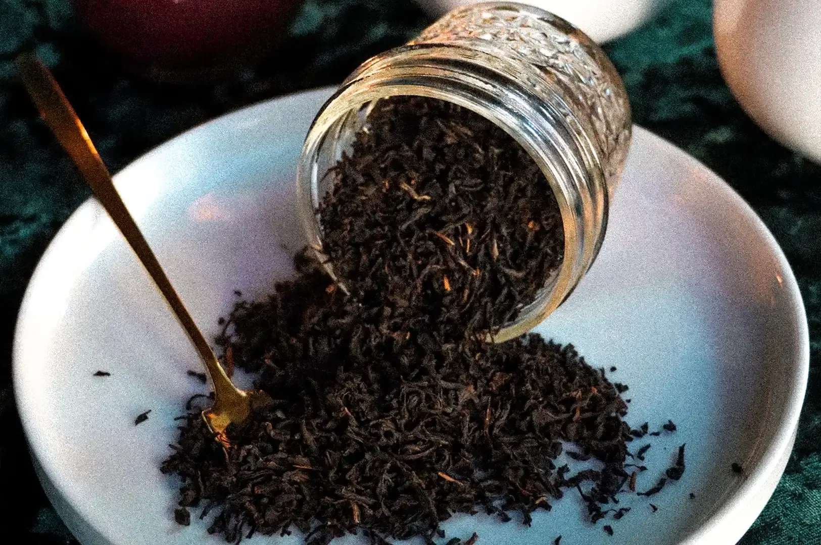 A stylized, moody composition showing a glass jar tipped over on a white plate, spilling dark Lapsang Souchong tea leaves onto the surface, accompanied by a single elegant gold spoon resting in the pile.