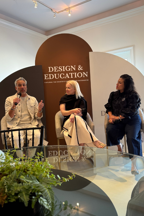 Three interior design professionals sit on a stage during a panel discussion beneath a backdrop that reads “Design & Education.”