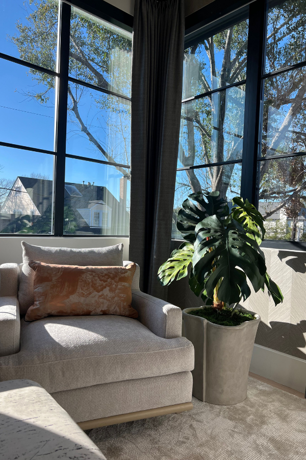Light gray armchair with a rust-colored pillow beside a large window and a potted monstera plant.