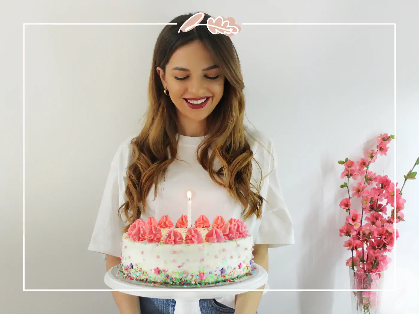 A happy woman with long hair holding a birthday cake with pink frosting and a single candle, standing beside a vase of pink flowers. Fabulous Flowers and Gifts.