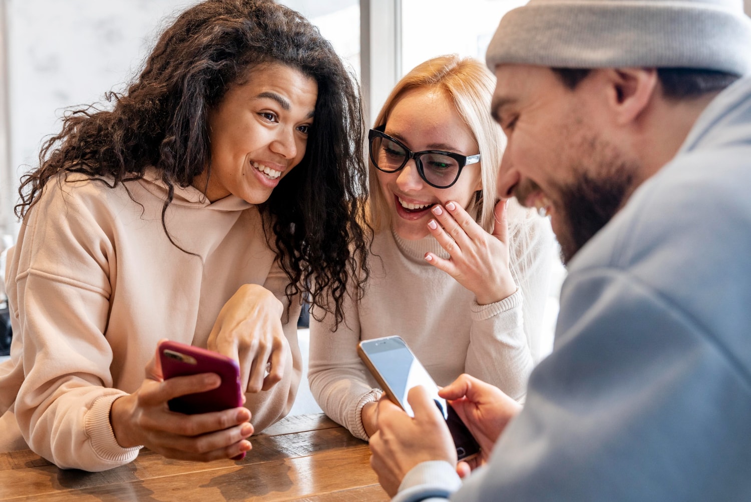 Three people gathered around a table enthusiastically looking at smartphones. Their energetic expressions suggest a lively discussion or shared discovery, demonstrating how mobile messaging solutions enhance collaborative internal business messaging.