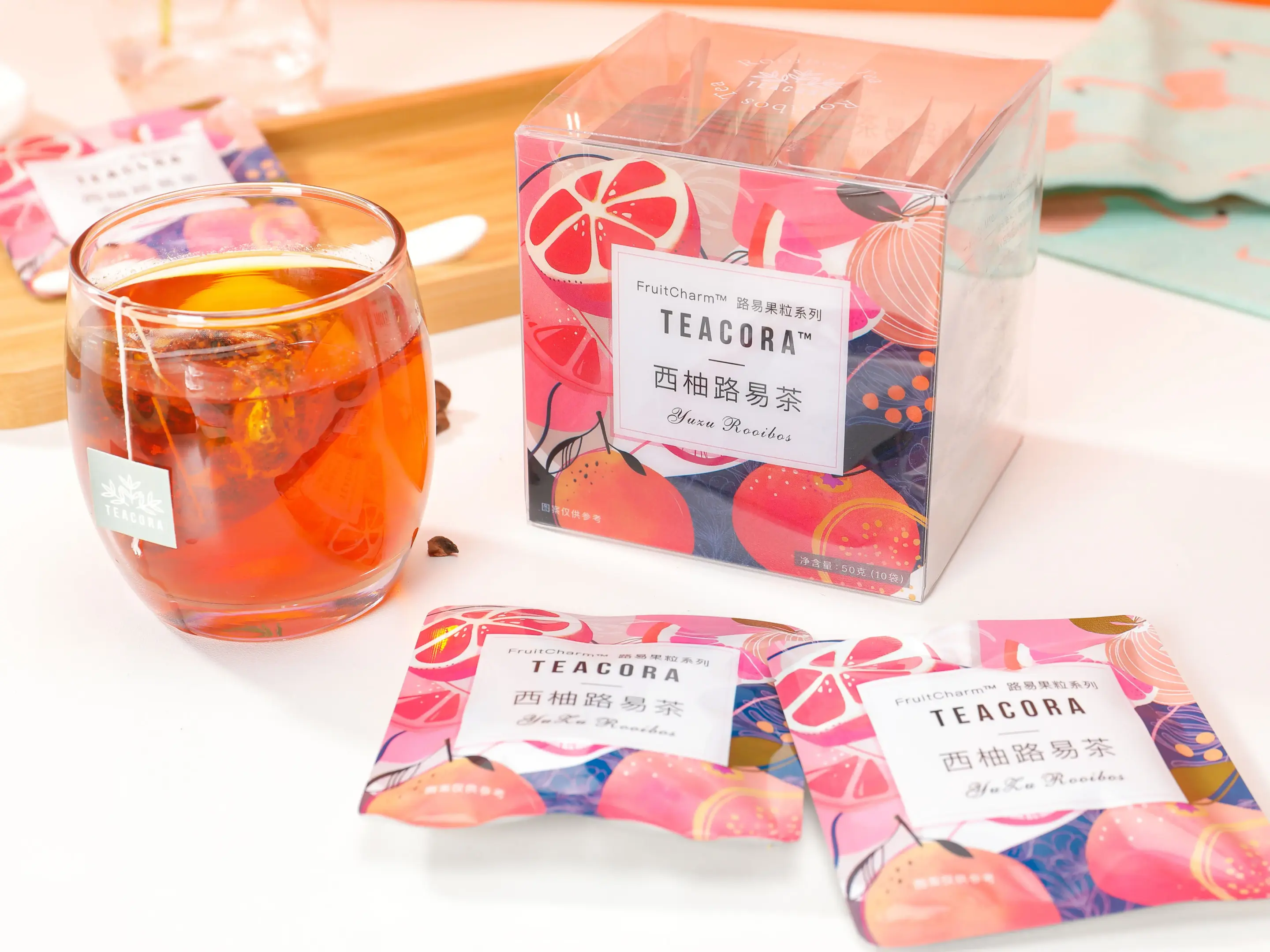 A clear glass cup of amber-colored tea sits alongside a decorative box and individual sachets of "Yuzu Rooibos" tea. The packaging features vibrant pink and orange illustrations of citrus fruits, creating a bright and refreshing aesthetic.