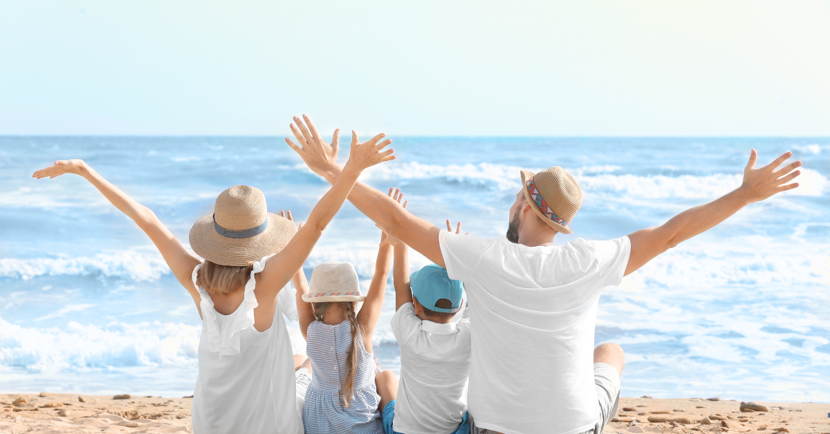 Family of four in sun hats sitting on the sand with arms raised, facing the ocean on a sunny day.