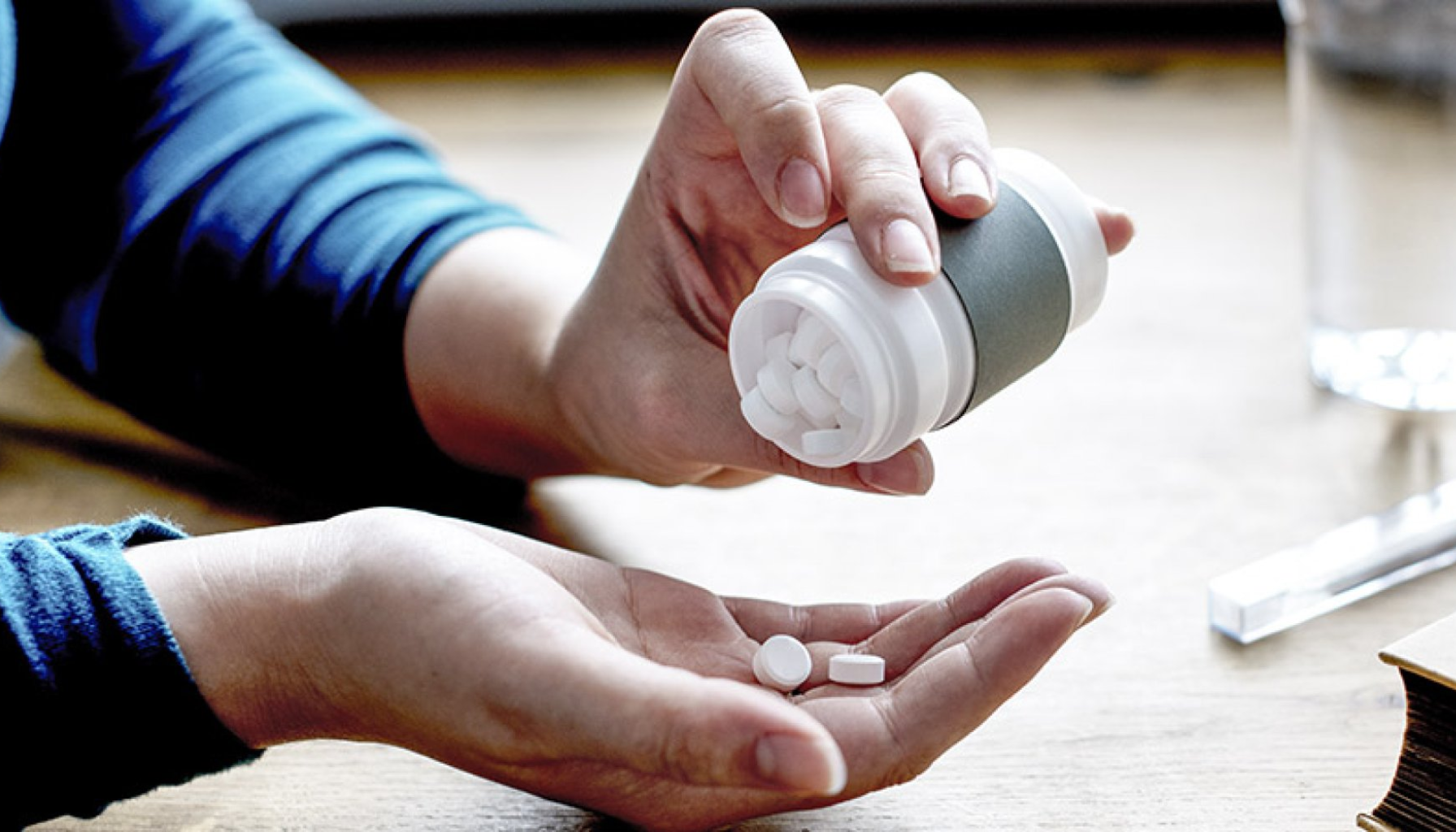 Person heller hvite tabletter fra pilleboks i hånden, klar til medisinering ved hjelp av vann som står ved siden av.