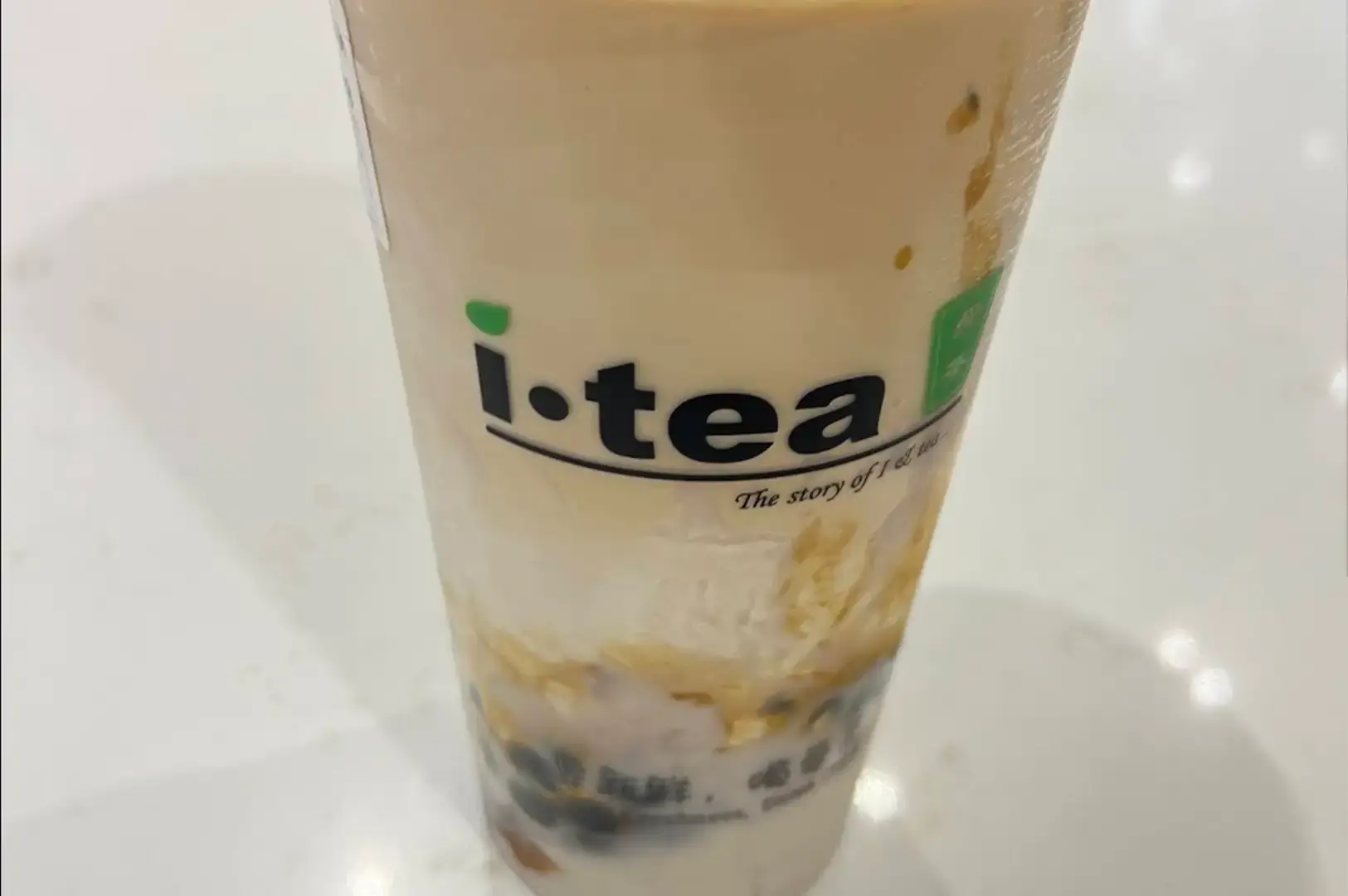 A close-up view of an iTea milk tea served in a clear plastic cup, showing distinct layers of creamy light brown tea over a rich bottom layer filled with dark tapioca pearls and pudding. The black "i-tea" logo with a green leaf accent is prominently displayed near the center of the cup.
