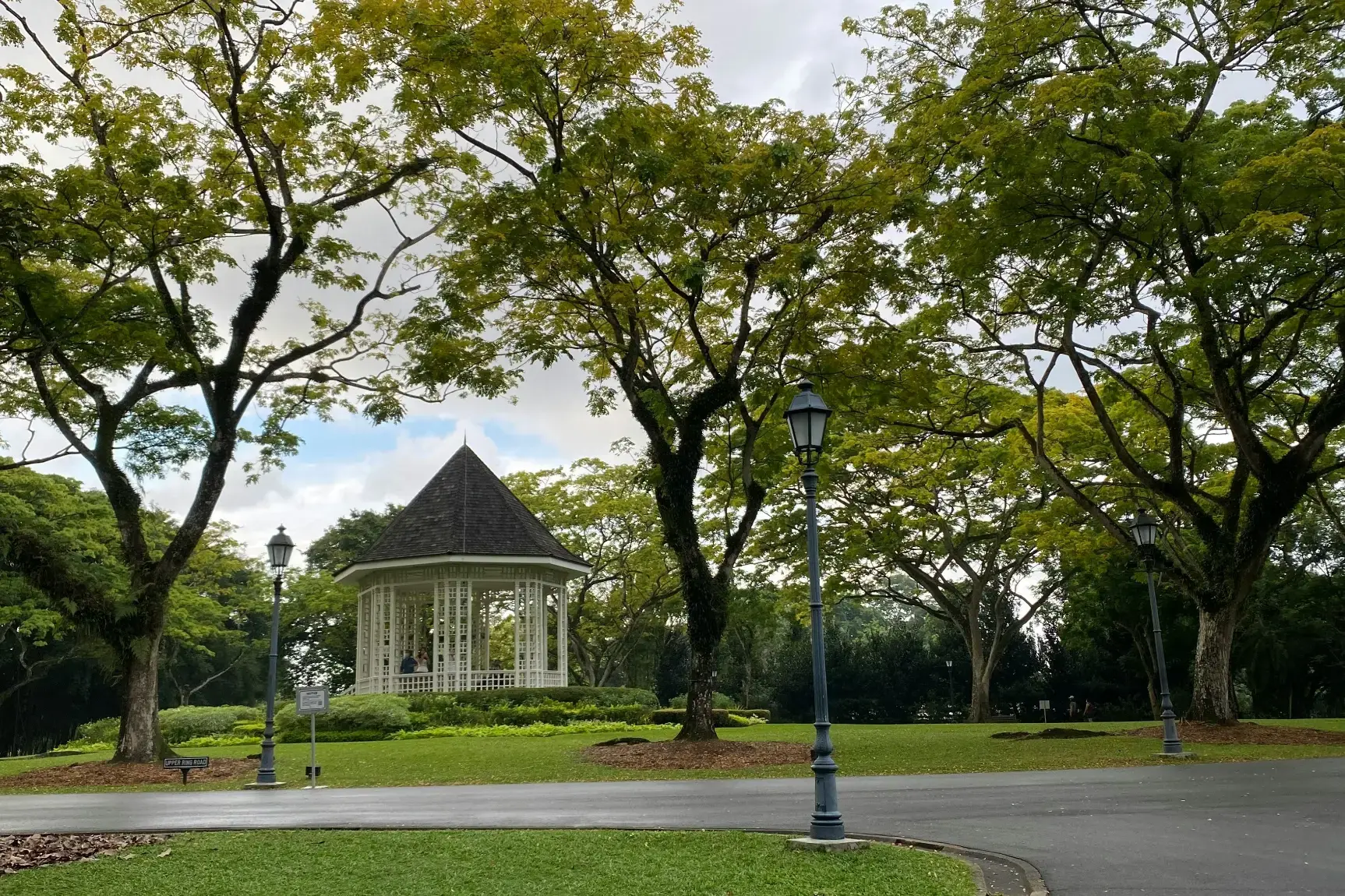 A white, octagonal gazebo with a dark conical roof sits nestled among lush green lawns and towering, leafy trees. Elegant vintage-style lamp posts line a paved path in the foreground under a soft, overcast sky.
