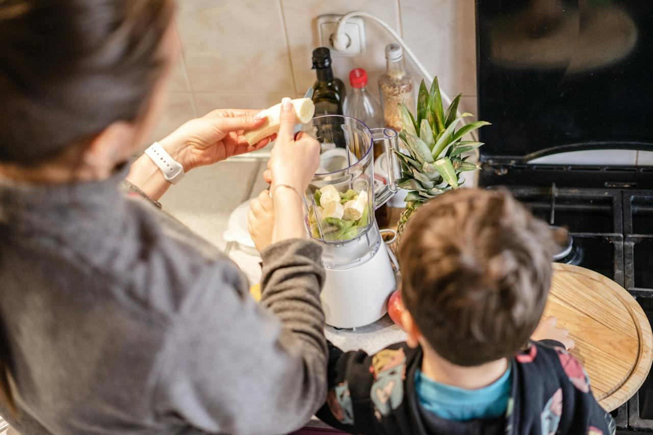 Uma pessoa adulta e uma criança estão juntas na cozinha, adicionando frutas em um liquidificador, preparando-se para fazer um suco ou vitamina.