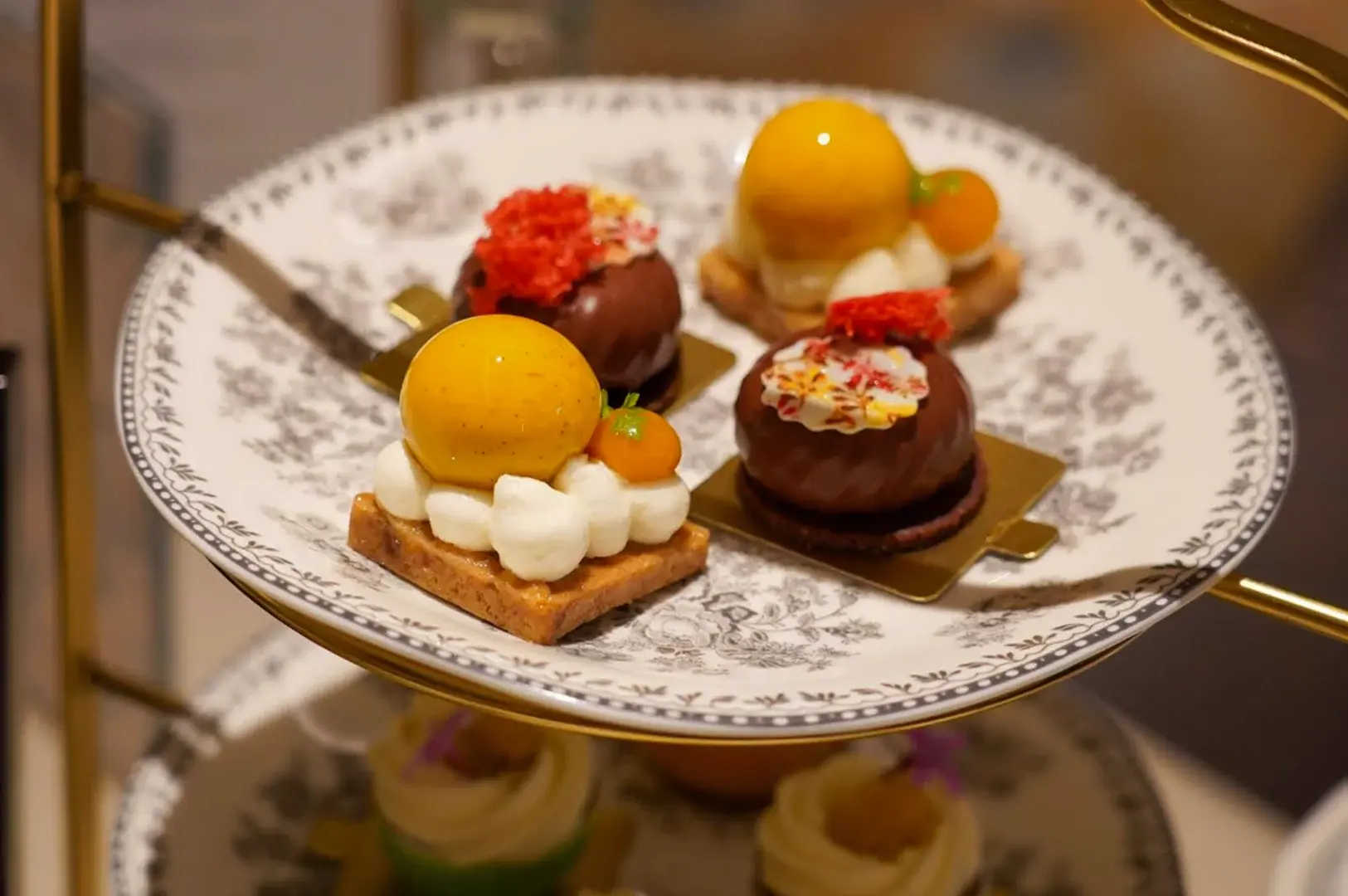 This close-up shot highlights the top tier of a golden afternoon tea stand, holding a white porcelain plate with a delicate black-and-white floral pattern that displays four gourmet desserts. Two of the pastries are square-shaped shortbread crusts layered with white cream dollops and topped with a vibrant, glossy yellow spherical glaze and a tiny orange garnish, while the other two are round chocolate domes resting on dark cookie bases, beautifully decorated with white cream, colorful sprinkles, and a tuft of red sponge cake garnish. The lower tier of the stand is visible in a soft blur below, showing additional creamy green pastries against a warm, neutral background.