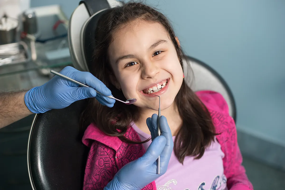 Smiling girl receives dental examination with instruments in dental chair.