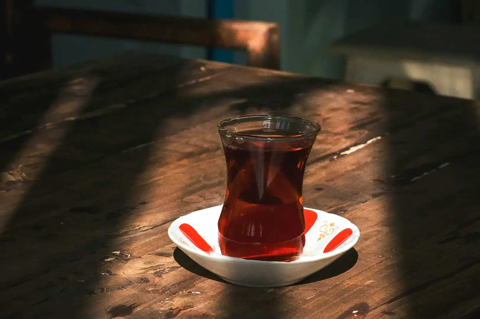 A single glass of rich, dark amber Turkish tea rests on a traditional white saucer with red geometric accents, placed in a beam of natural sunlight cutting across a dark, weathered wooden tabletop.