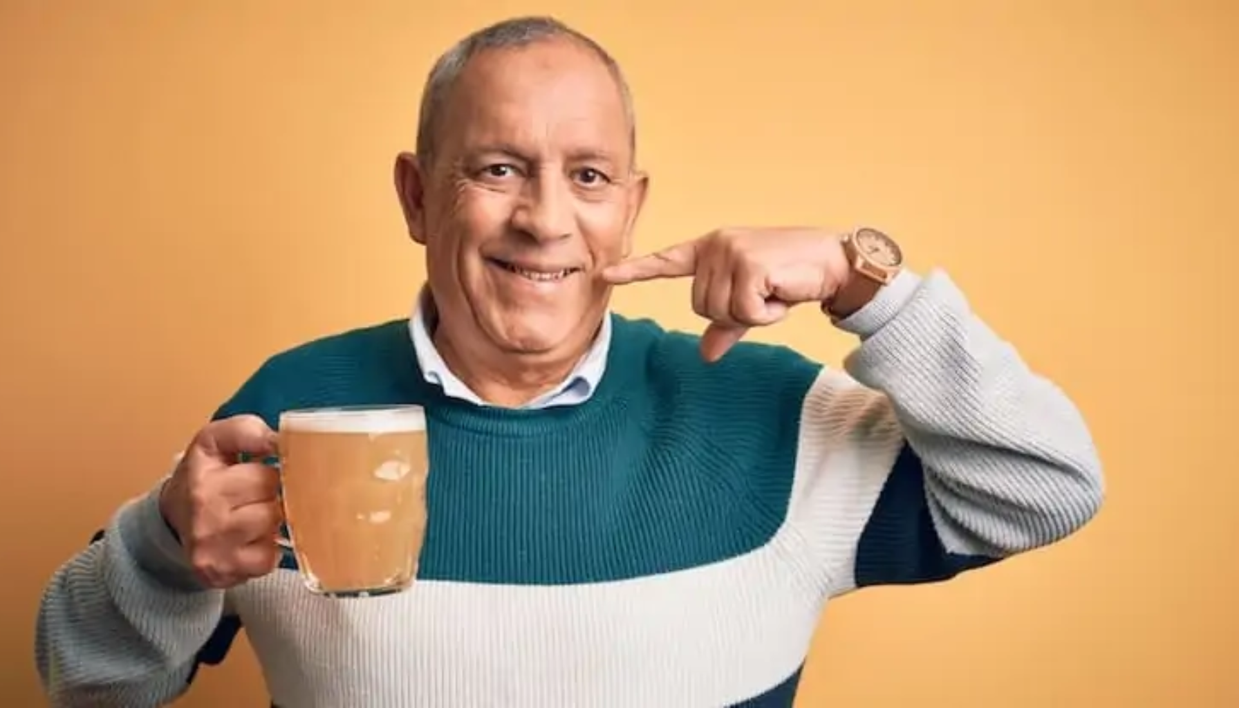 Smilende eldre mann holder en kopp kaffe og peker på tannen med fingeren foran oransje bakgrunn.