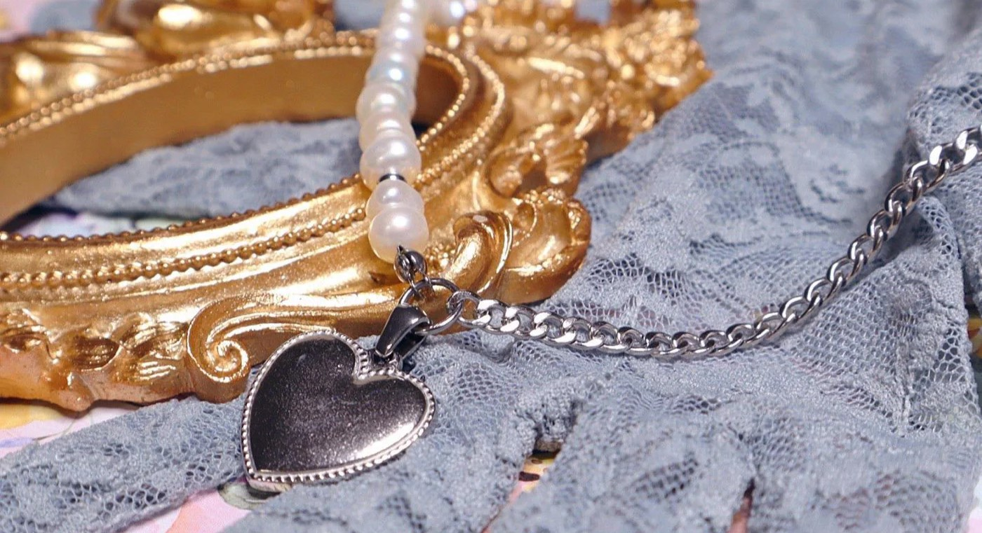 Close-up of a silver necklace with half pearls, half curb chain, featuring a heart pendant, laid over a blue lace-patterned glove and beside a gold picture frame.