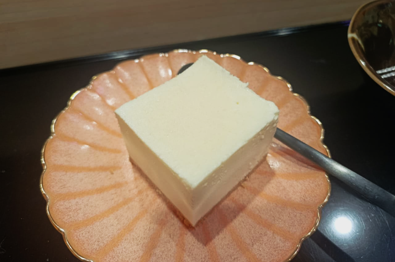 Square slice of white tofu on a round, pink scalloped plate, with a metallic spoon beside it. The setting is clean and minimal.