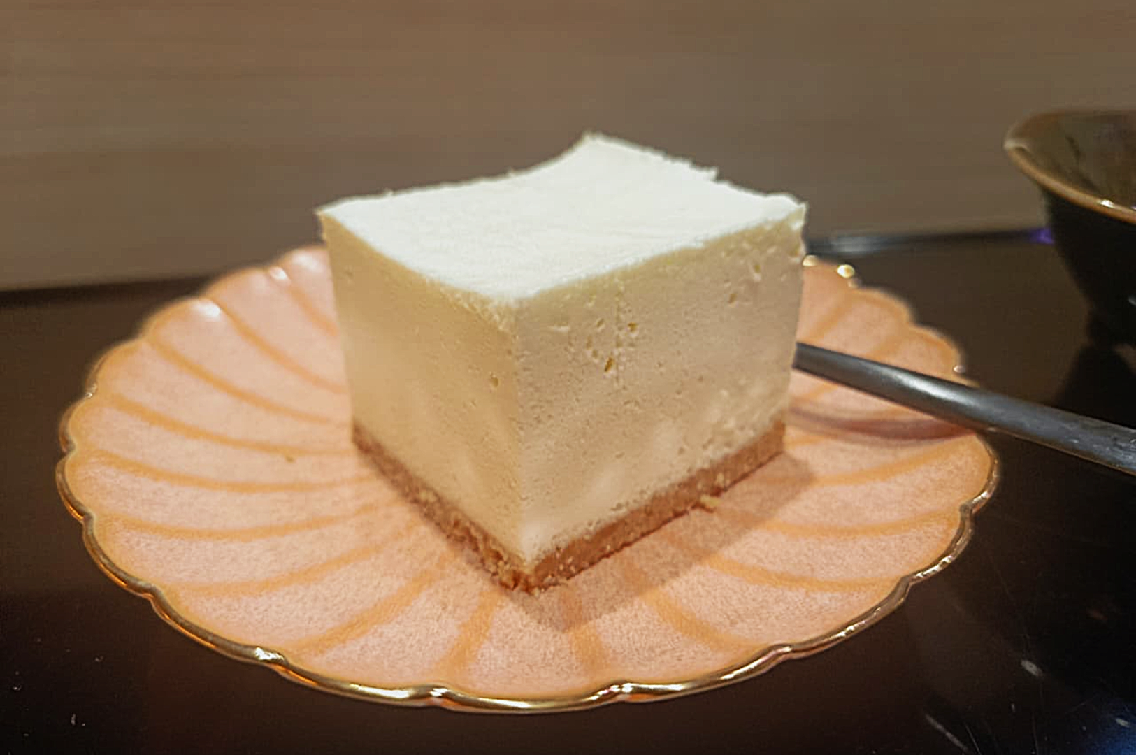 A square slice of creamy cheesecake with a light brown crust sits on a pink, scalloped plate. A spoon is placed beside it, suggesting readiness to eat.