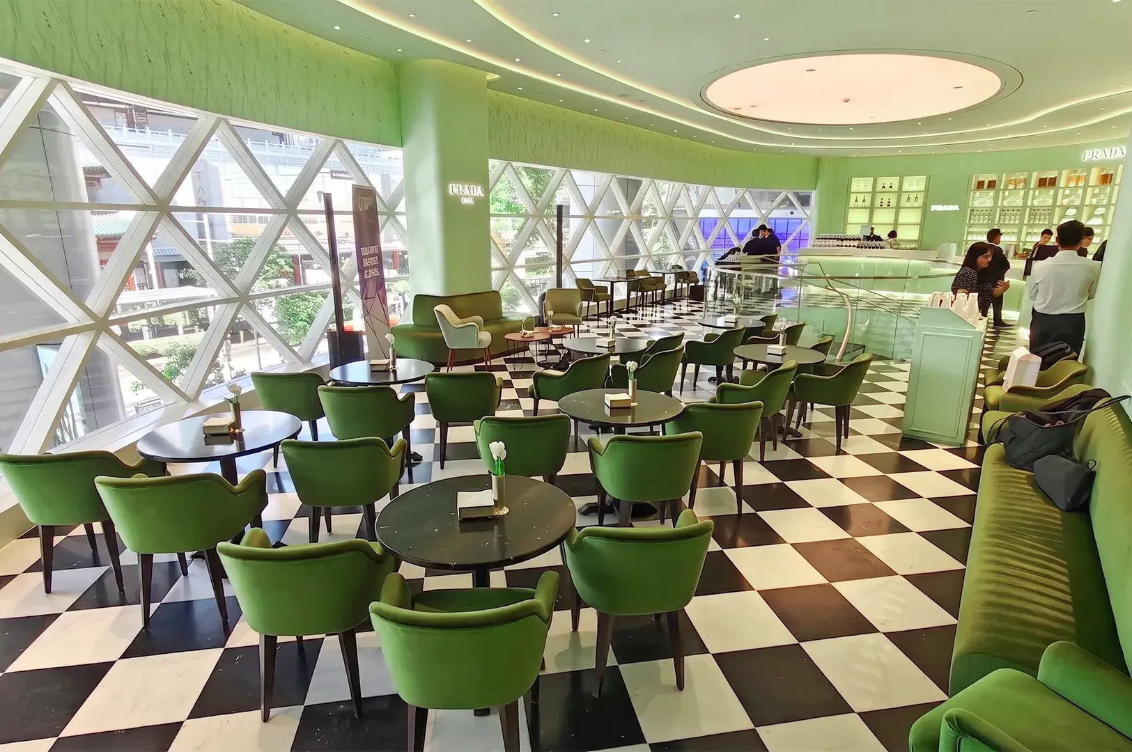 Spacious Prada Cafe seating hall featuring mint green armchairs, dark round tables, checkered flooring, and large geometric lattice windows.