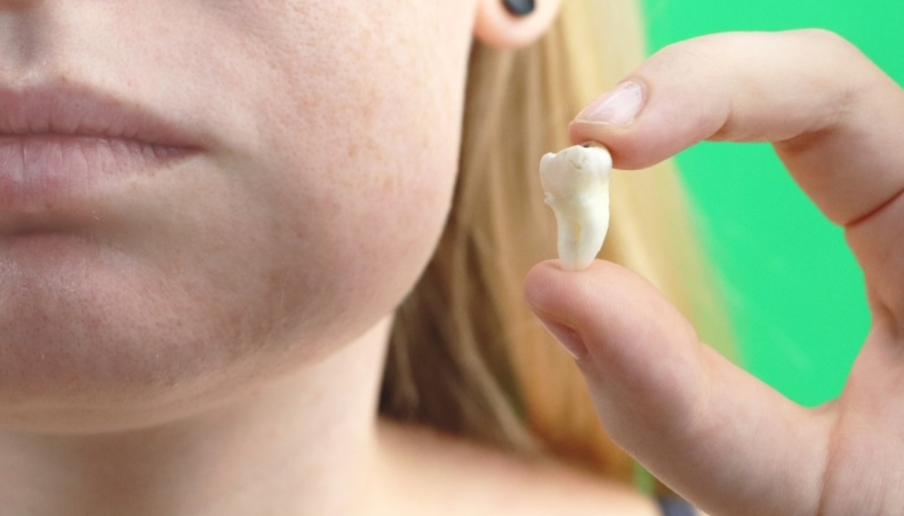 Betennelse Etter Trekking av Tann: Når Helingen Går Galt 12 Nærbilde av person som holder en trukket tann mellom fingrene, med nøytralt ansiktsuttrykk og grønn bakgrunn i studio