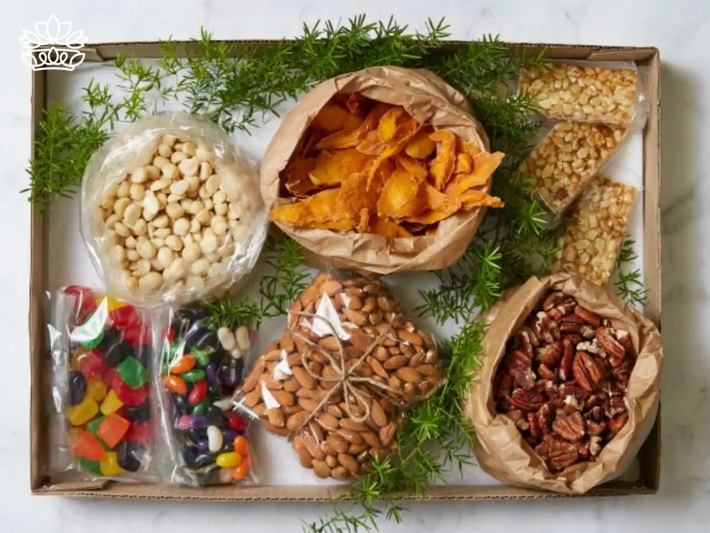 An assortment of gourmet snacks in a gift box, featuring macadamia nuts, dried mango, assorted seed bars, almonds, pecans, and colorful candies, garnished with fresh green herbs. Fabulous Flowers and Gifts.
