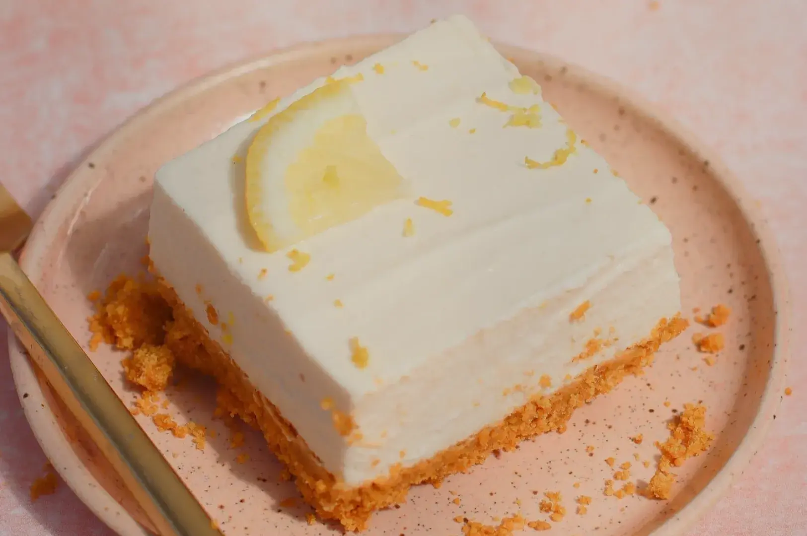 A square slice of lemon cheesecake on a plate, topped with a lemon slice and some lemon zest. Crumbles of the crust are scattered around the plate.