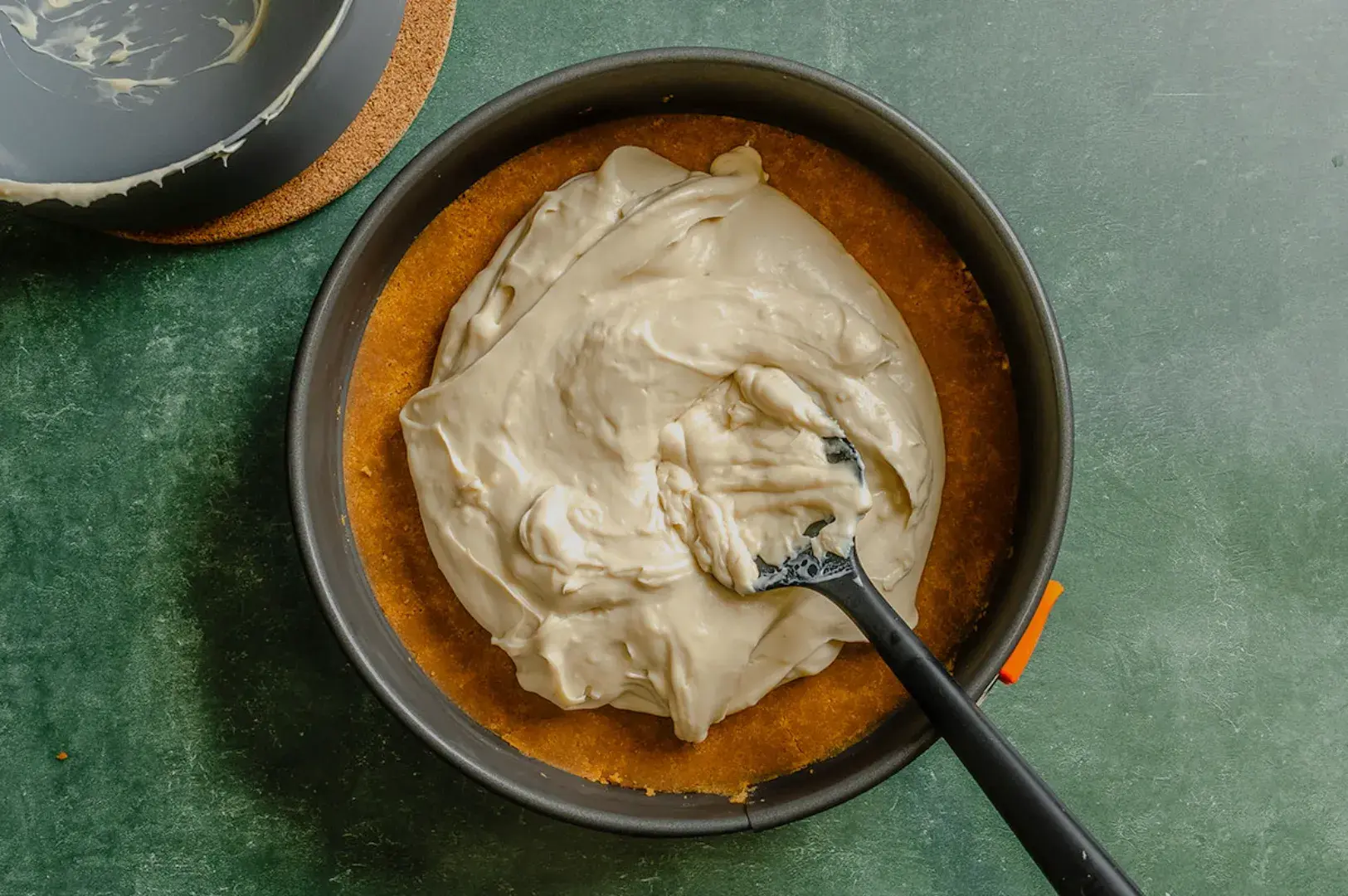 A round cake pan with a graham cracker crust topped with a creamy filling and a spatula resting on top.