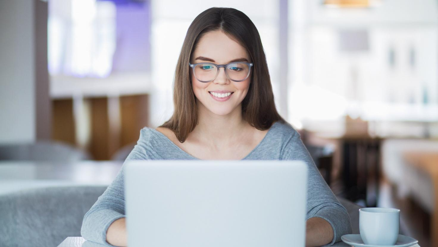 A young woman wearing glasses smiles while working on a laptop in a bright, modern space. This image conveys individual participation in virtual meetings, showcasing how video conferencing enables remote collaboration from any location.