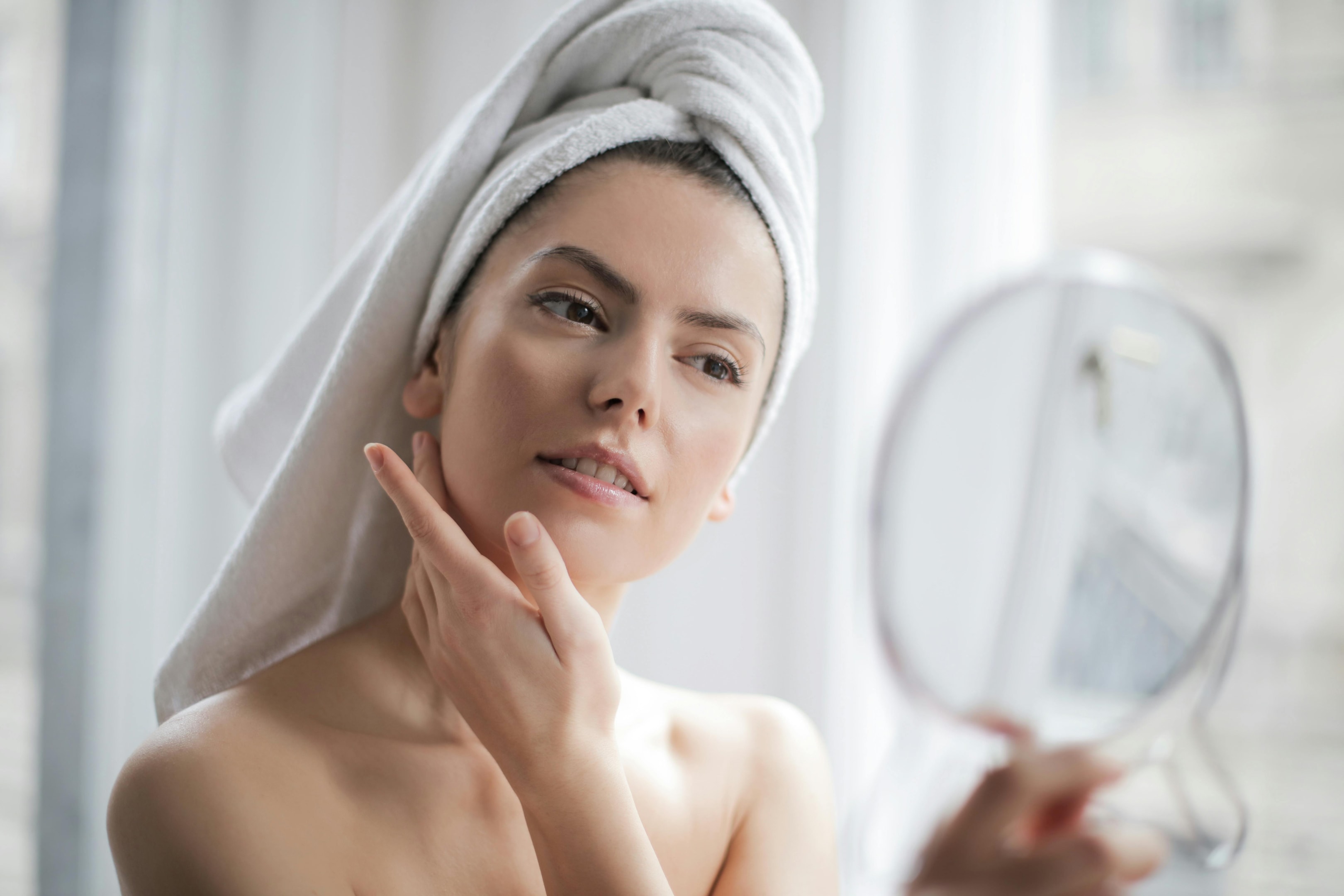 A woman with a towel wrapped around her head looking at her reflection in a hand mirror. She gently touches her face as if she's just taking care of her skin or preparing for the morning. The scene is calm and serene, with soft lighting highlighting her natural complexion.