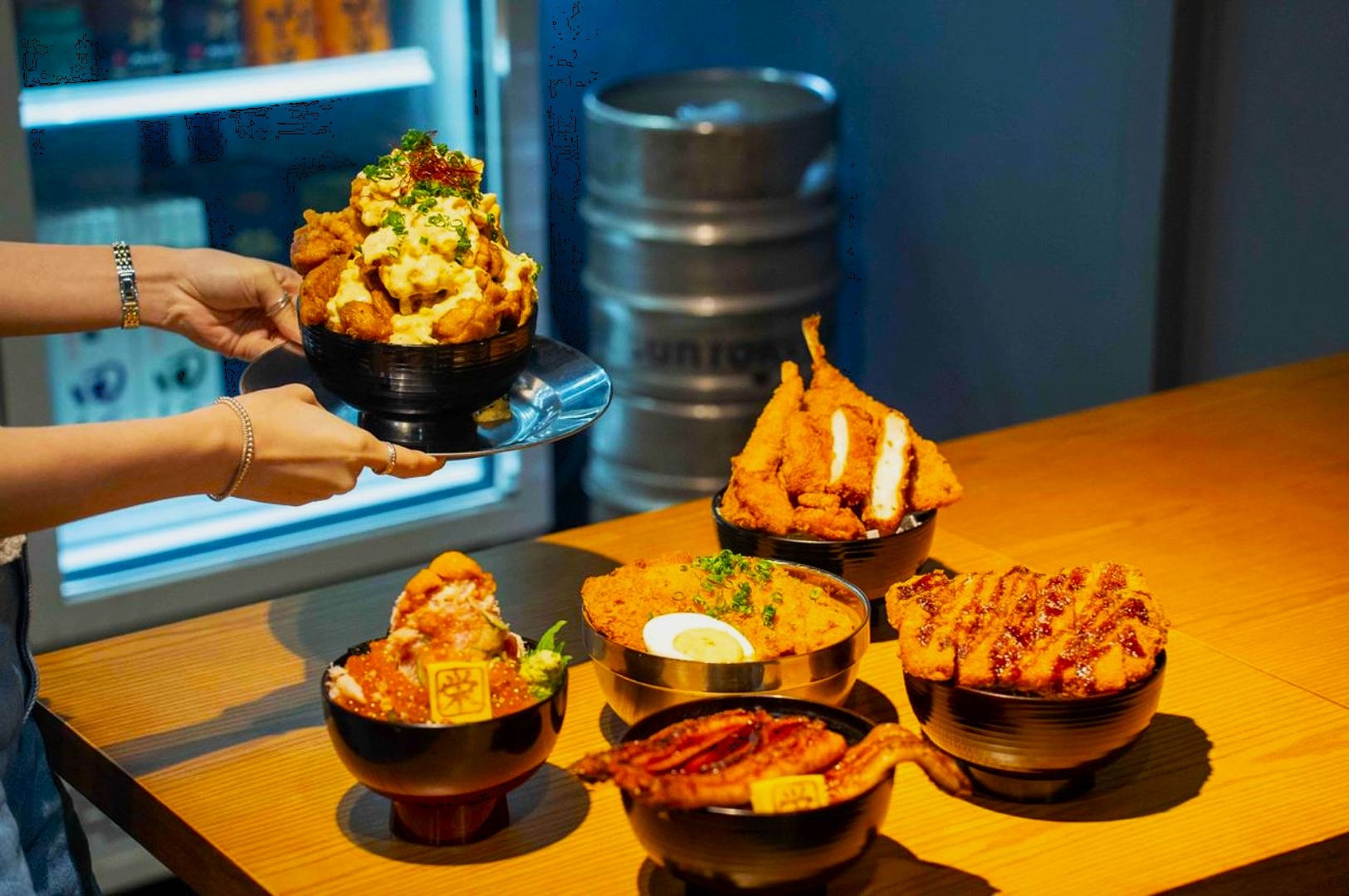 A variety of Japanese rice bowls on a wooden table, featuring fried chicken, grilled eel, and cutlets. A hand serves a bowl topped with creamy chicken.