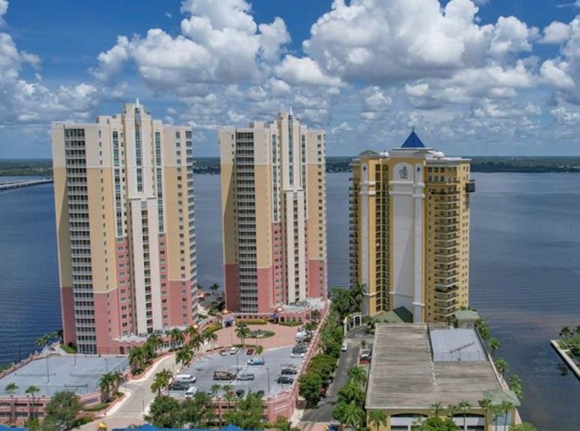 An aerial view captures the vibrant downtown waterfront district of Fort Myers, showcasing high-rise buildings and a bustling marina. The image highlights the stunning sunset views over the Caloosahatchee River, a backdrop for the luxurious Beau Rivage condos, offering a truly remarkable living experience.
