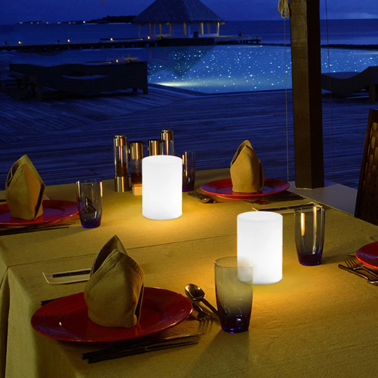 Outdoor dining table setup with glowing LED candles, folded napkins, and red plates overlooking a pool and ocean view at night.