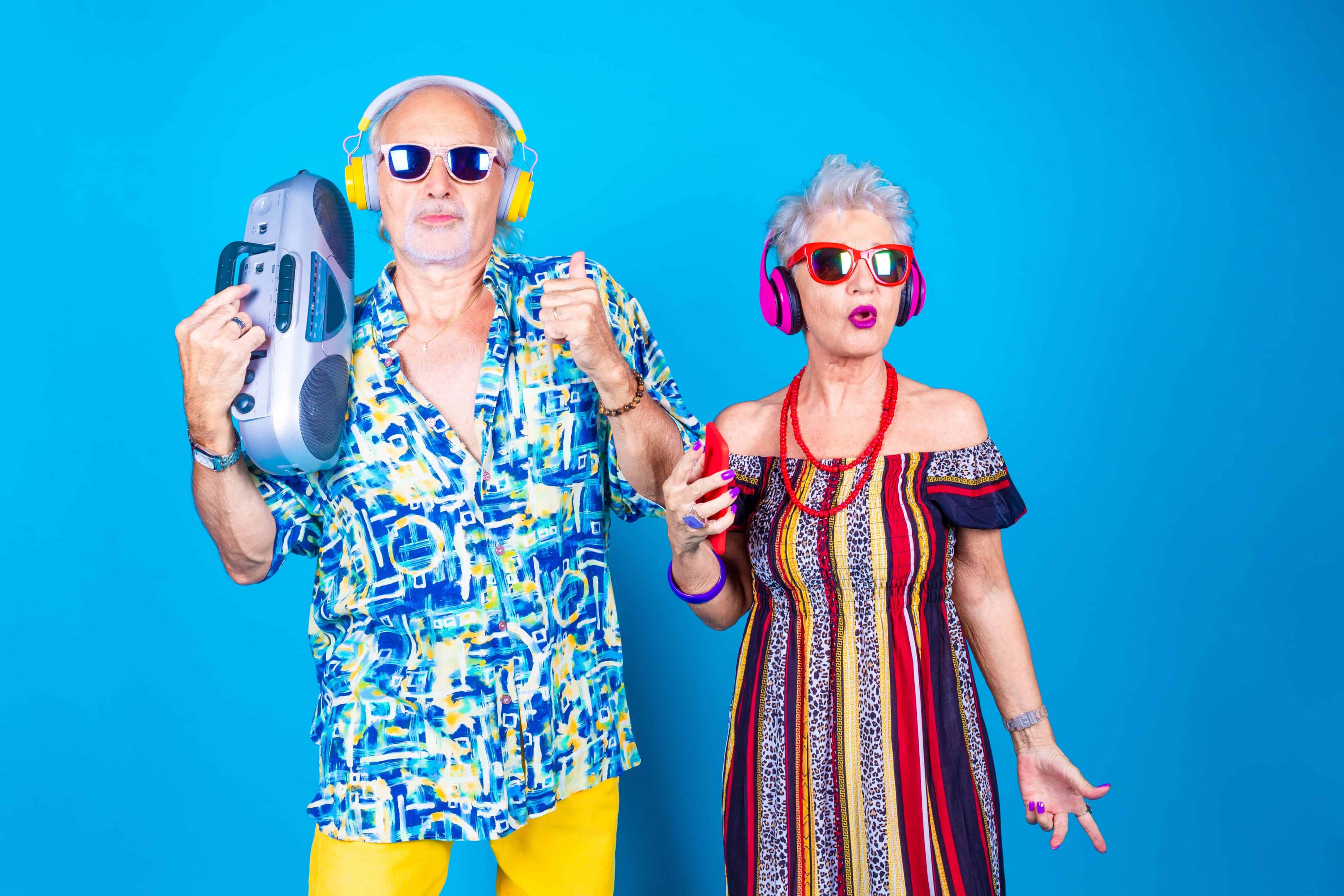 An older guy with a healthy body in glasses and a boombox. An older woman with glasses and jewelry on. Together they are enjoying the music and and dancing.