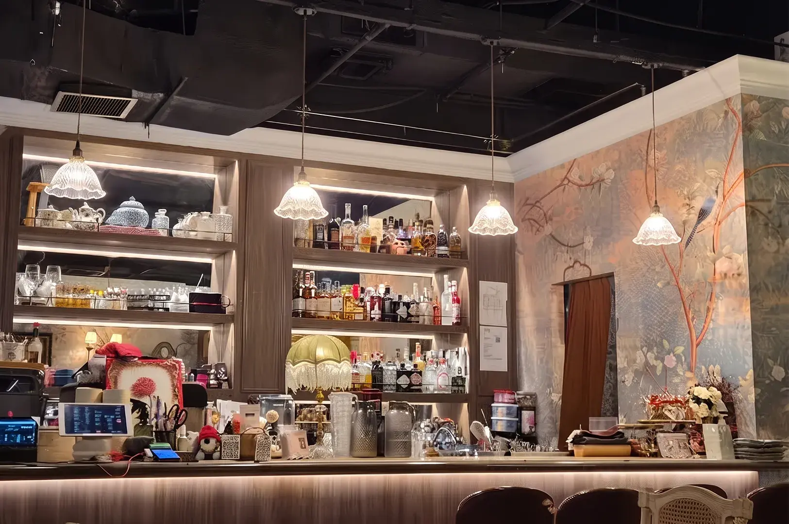 Elegant cafe and bar counter featuring warm backlit shelving stocked with spirits and teapots, delicate scalloped glass pendant lights, and a floral wallpaper accent wall.