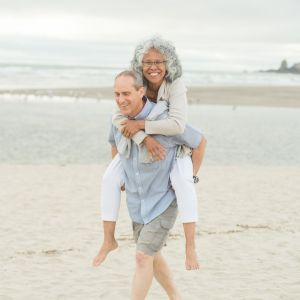 older couple walking on the beach and the man is carrying the woman piggy back