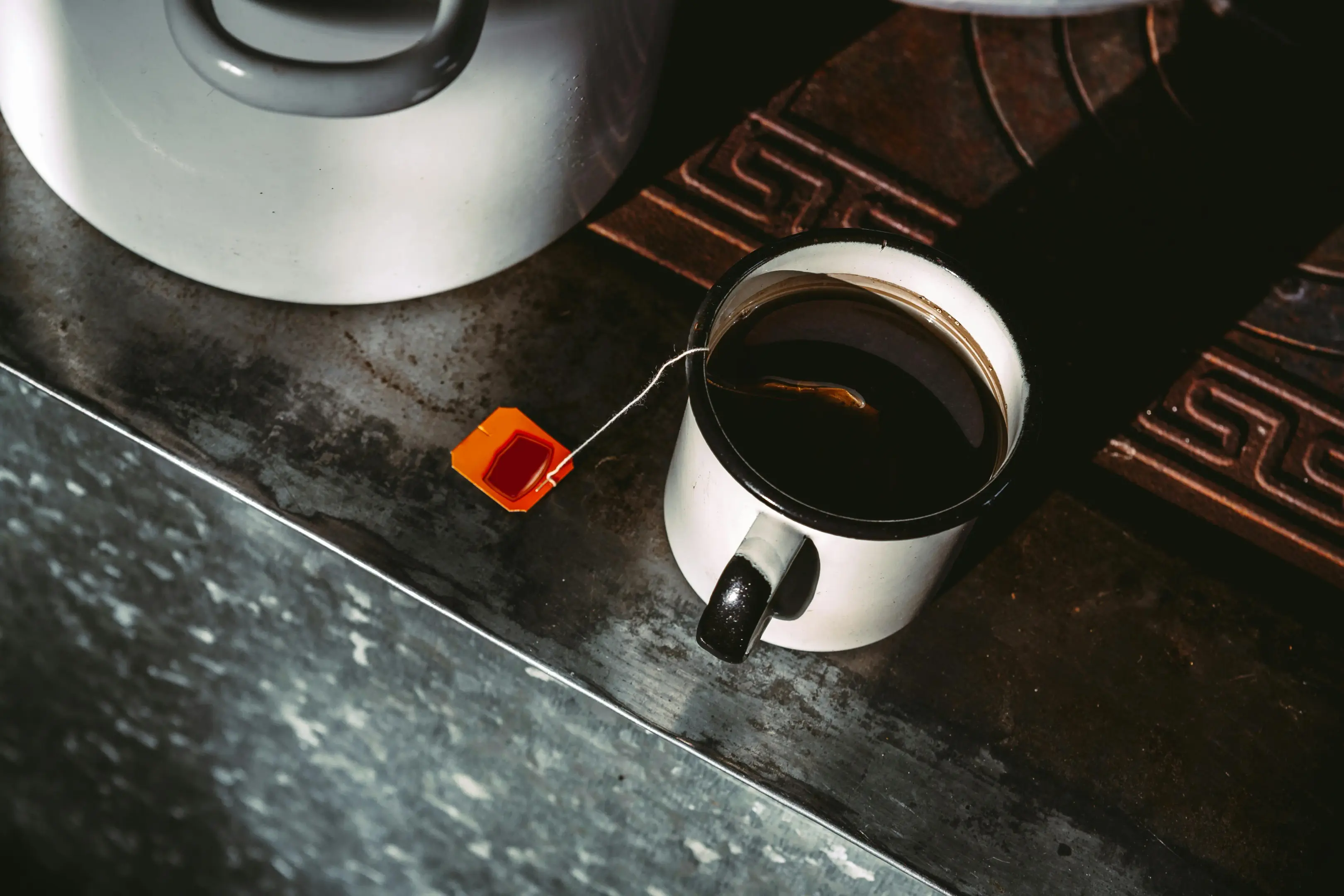 A high-angle shot shows a white enamel mug filled with dark tea, its orange teabag string draped over the side onto a rustic, weathered surface. Next to the mug sits a large white pot, all illuminated by a dramatic, sharp shadow slicing across the scene.