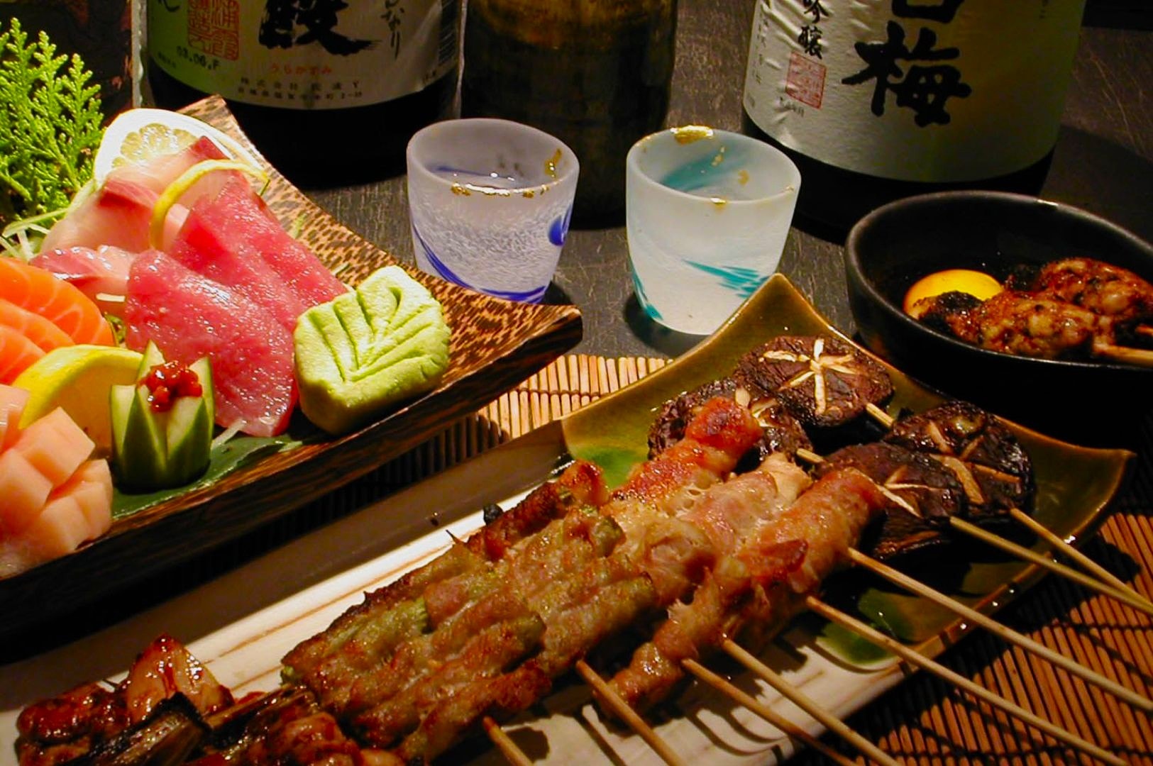 A colorful Japanese meal with sushi, sashimi, and yakitori skewers on platters. Drinks in elegant glasses, with sake bottles in the background.