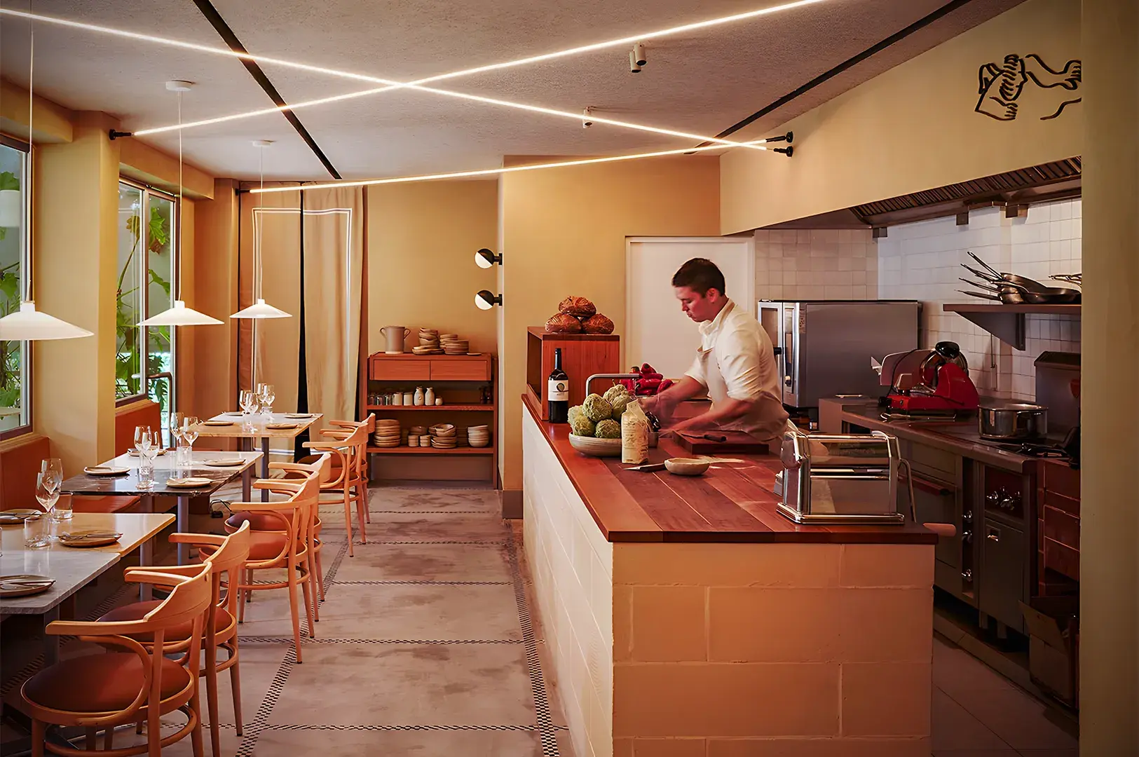 A warm, modern restaurant interior with wooden tables and chairs. A chef prepares food at a counter, with bright lights and an organized kitchen.
