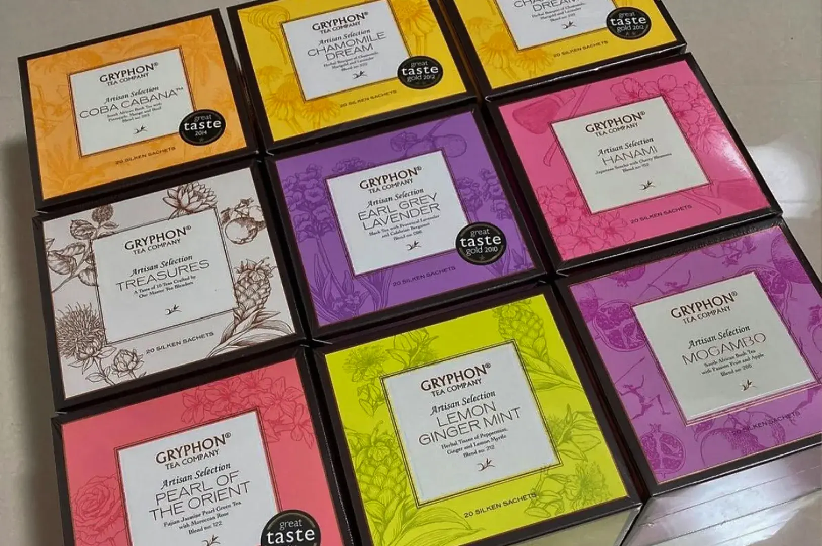 An overhead grid arrangement of nine distinct, colorful Gryphon Tea Company boxes from their Artisan Selection line laid out flat next to one another. The collection showcases a variety of flavors with color-coded packaging: top row features orange "Coba Cabana" and yellow "Chamomile Dream"; middle row contains a white "Treasures" box, a purple "Earl Grey Lavender" box with a "Great Taste" award sticker, and a bright pink "Hanami" box; bottom row includes a salmon-pink "Pearl of the Orient," a lime-green "Lemon Ginger Mint," and a deep purple "Mogambo" box. Each individual square box displays the brand name, blend title, and unique floral or botanical line art graphics reflecting its ingredients.