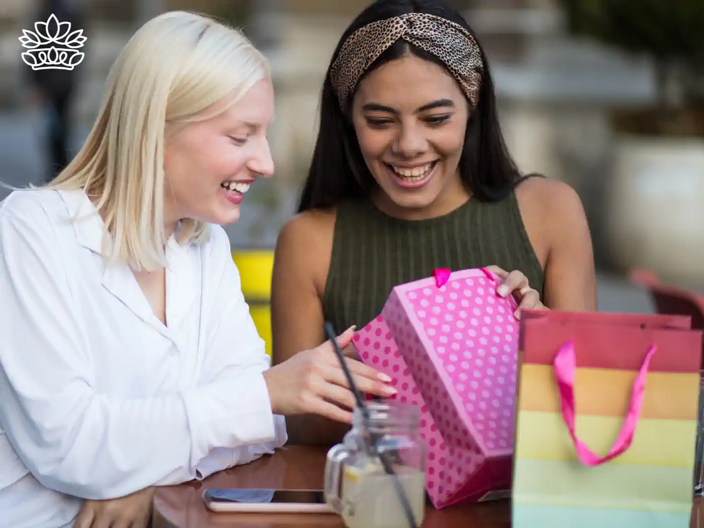 Two joyful women sharing a lively moment while looking at colorful gift bags at an outdoor café. Fabulous Flowers and Gifts - Thank You Gift Boxes. Delivered with Heart.